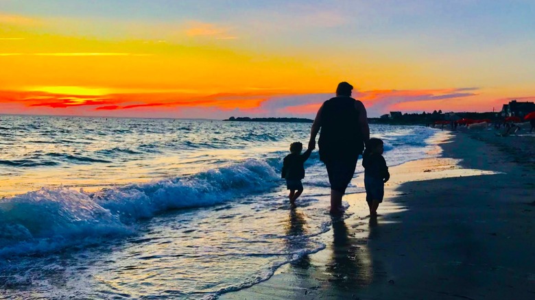 Silhouettes of a parent walking hand-in-hand with two children along Old Silver Beach at sunset, with a blazing orange sunset