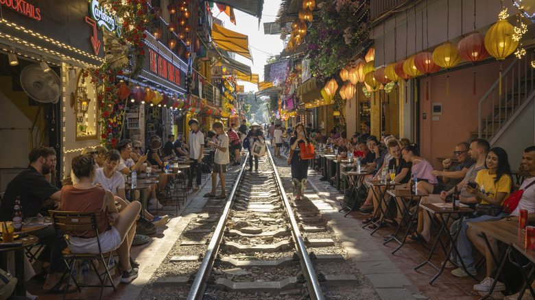 Tourists sitting on restaurant tables under colorful lanterns and neon signs along a railway track in Hanoi