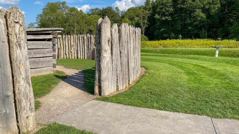 Reconstructed wooden structures of Fort Necessity, with a circular wooden wall on the left surrounded by grass, with trees in the background under a blue sky