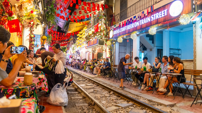Diners seated on tables along both sides of a train track with neon lights and red flags overhead