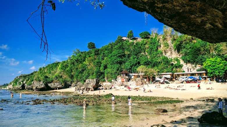 Rugged jungle cliffs stand above a tourist filled beach at low tide.