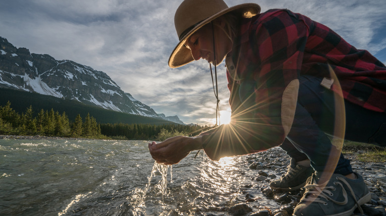 A woman crouches before a river at sunset and gathers water in her cupped hands