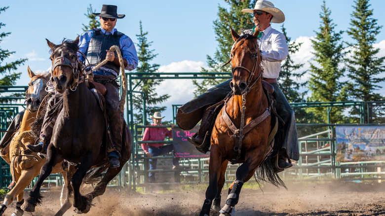 Two men in cowboy outfits ride around a dirt arena, raising dust