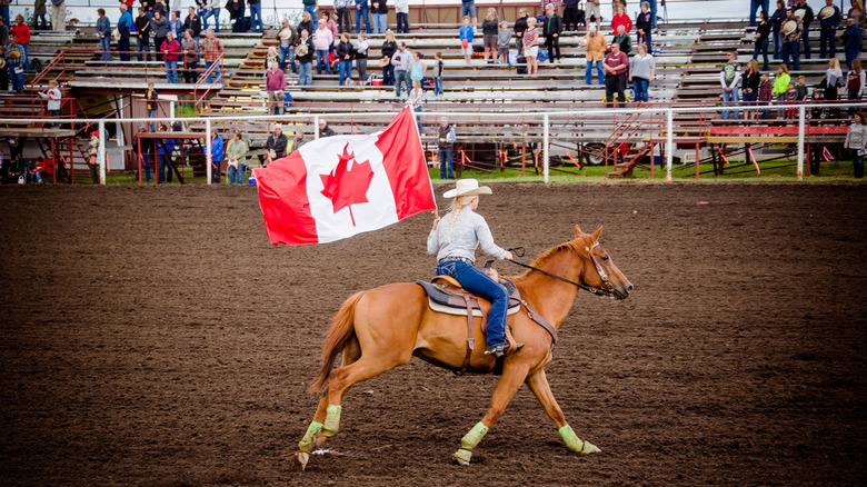 A woman rides a horse around a rodeo arena while carrying the national flag of Canada