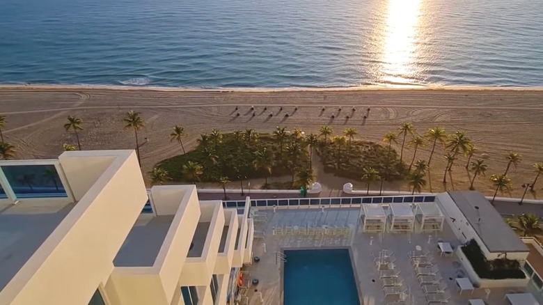 Tall angular white hotel and pool across from beach flanked by coconut palms.