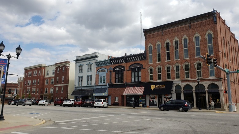 Row of brick commercial buildings, one with a white-painted facade, with cars parked along a street out front under a cloudy sky