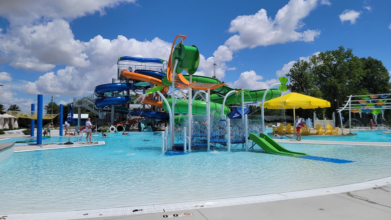 Water park, with a green and orange playground and yellow beach umbrella in the foreground of blue, orange, and green water slides, under a cloudy blue sky