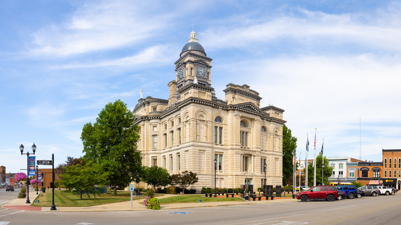 Courthouse in Frankfort, Indiana, a white stone building with a dome surrounded by trees, with cars parked beside it and downtown shops in the background, under a cloudy blue sky
