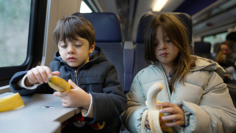 Children sitting side-by-side on a train peeling bananas