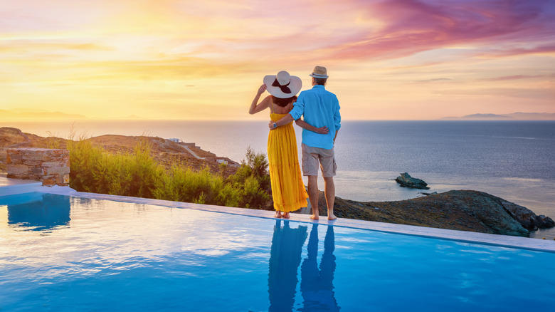 A couple standing along a pool with a view of the ocean beyond. Their backs are turned as they watch a colorful sunset transform the sky above them.