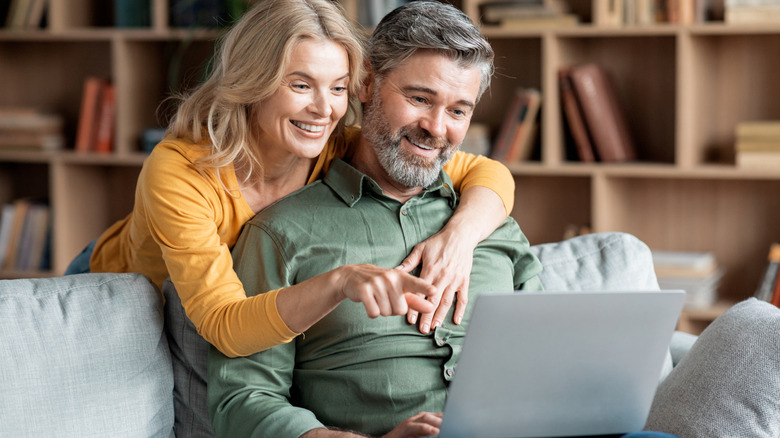A middle-aged couple looks at the screen of a laptop together. The man is sitting on a comfortable sofa while the woman stands behind him, smiling.