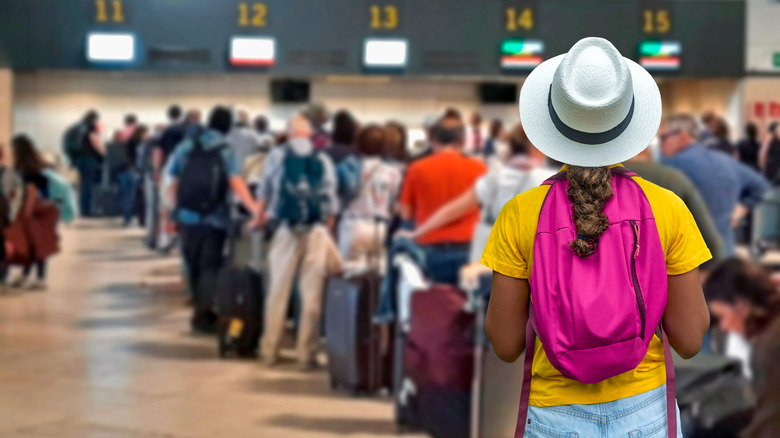 A tourist girl in a yellow shirt, pink backpack, and white hat waits in a long line at an unidentified airport with other passengers to check her luggage