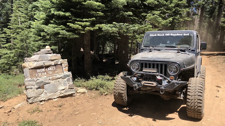 A dusty off road vehicle (Jeep) sits next to the iron lakes trailhead sign reading Iron Lakes 23E04 1 1/2 miles with a right facing arrow; the background has vibrant green trees and red-tinged dusty roads