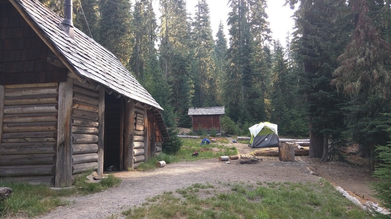 Wooden shelter with a small red-walled wooden shack and gray-and-green tent in the background