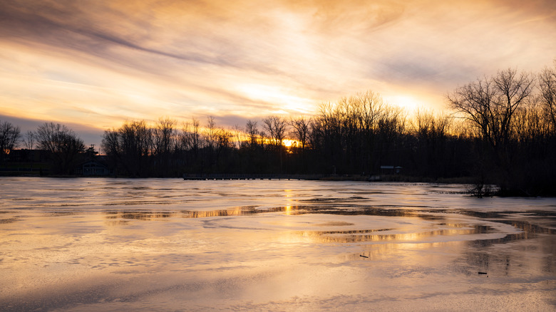 Sun setting over a partially frozen Reeds Lake in East Grand Rapids, Michigan, with leafless trees lining the shore
