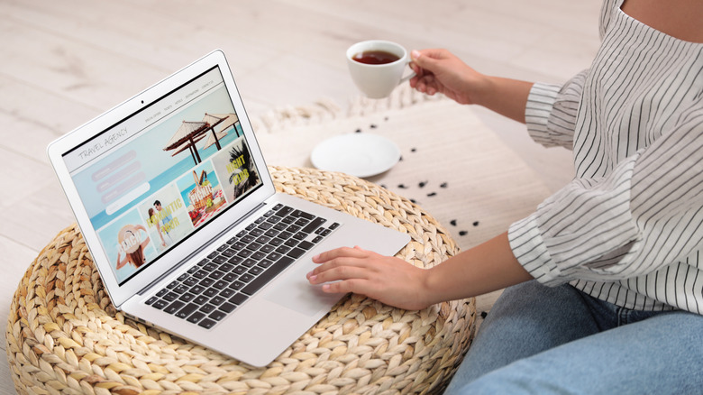 Woman holding cup of tea and looking at a travel agency webpage on her laptop
