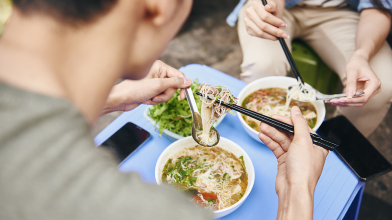 Two people eating bowls of Asian noodles with chopsticks and spoons