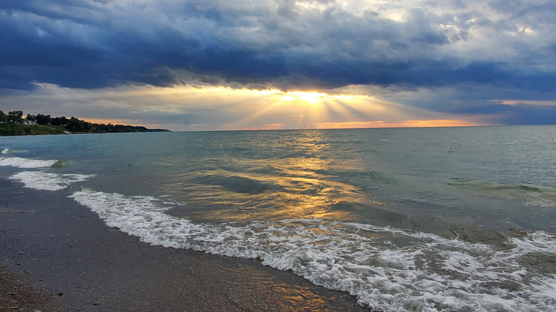 Conneaut Township Park at sunset, with white surf in the foreground, a treed shore on the left side of the frame, and sunbeams from a cloudy sky in center frame