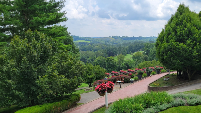 Red path beside trees, with pink flowers hanging from a white post beside it, leading to a wooden bench and a trellis with red flowers, with green treed hills in the background, under a cloudy sky