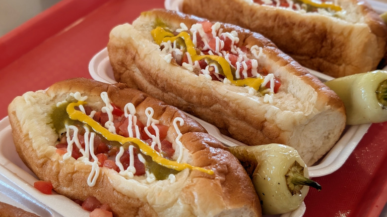 Three Sonoran hot dogs with condiments and peppers on red tray