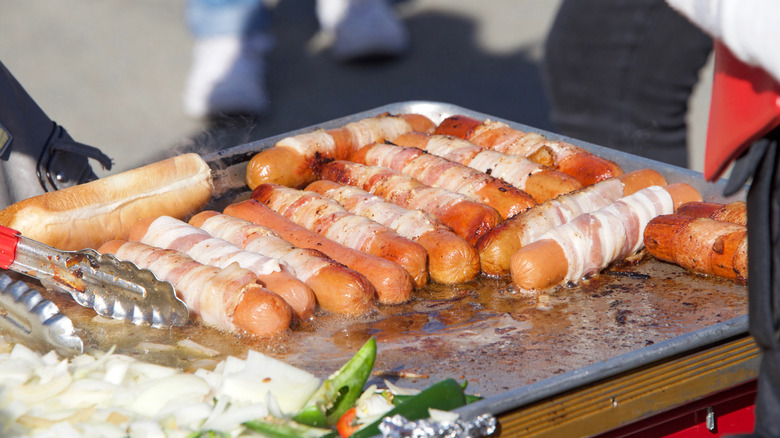 Bacon-warpped hot dogs on grill with onions and tongs