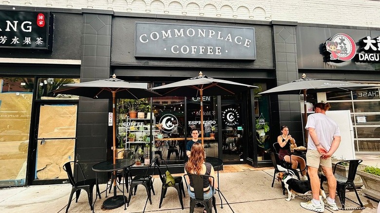 Exterior of the Squirrel Hill Commonplace Coffee, with a black sign on a black wall and people sitting at black tables under black umbrellas with wooden poles on a concrete sidewalk