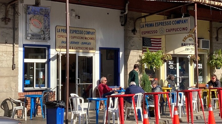 Front of La Prima, with people sitting at red, blue, and yellow tables surrounded by white chairs in front of white and yellow walls with signs