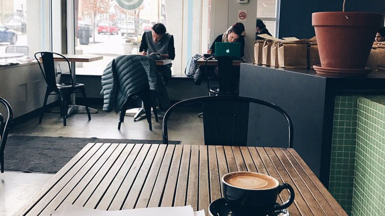 Wooden table with a black mug of coffee in the foreground, with people studying in the background and a black and green-tiled counter lined in coffee bags along the right side