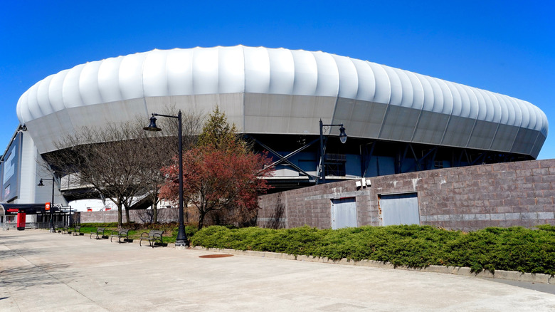 Sports Illustrated Stadium, a large white building behind trees next to a wide sidewalk under a blue sky