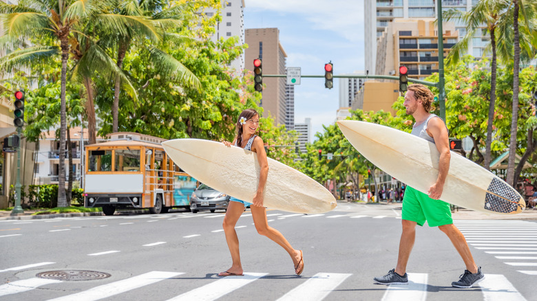 A young couple holding surfboards crossing palm tree-lined boulvard in Waikiki