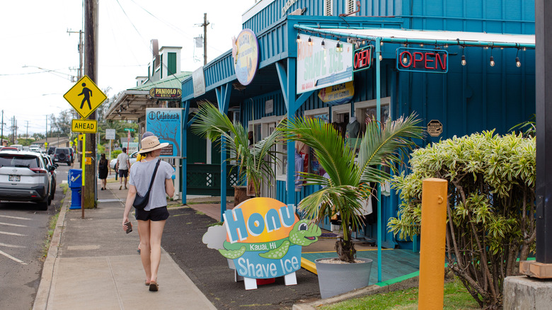 Female pedestrian walking along a sidewalk next to a blue shop and a shave ice sign
