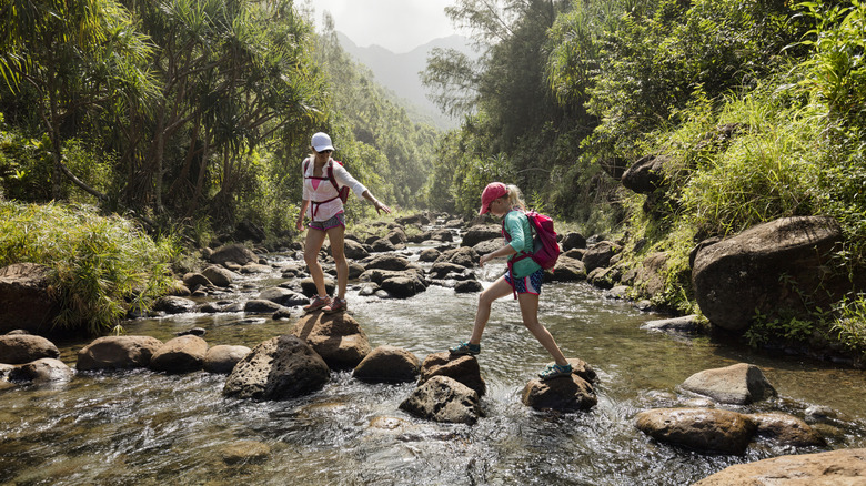 A mother and daughter crossing a creek in Nā Pali Coast State Wilderness Park in Kauai surrounded by leafy trees in Hawaii.