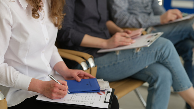 A row of seated people filling in forms while holding passports