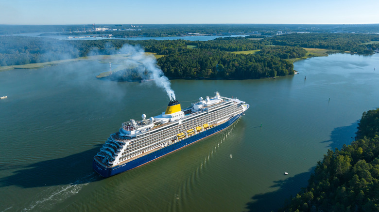 A large cruise ship going down a narrow stretch of water past trees and grass
