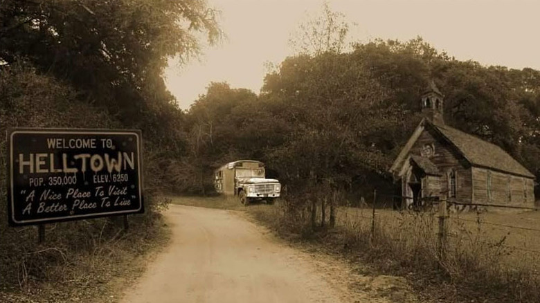 Sepia-tinted photo of a sign reading "Welcome to Helltown" beside a dirt road, with an old church to the right and an abandoned schoolbus straight ahead, surrounded by trees