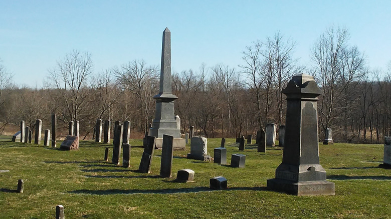 Boston Cemetery, with rows of old graves around a gray obelisk surrounded by grass, with leafless trees in the background under a blue sky