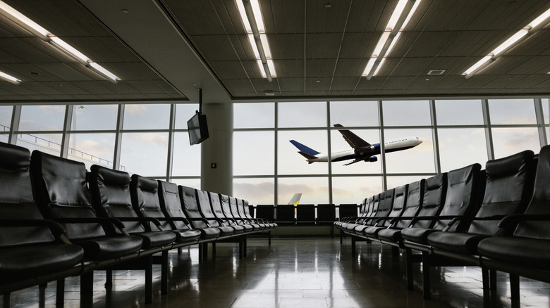 Two rows of empty airport terminal seats lead to a termnal window with a plane talking off