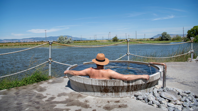 A man in a hat soaks in a hot spring tub overlooking a river.