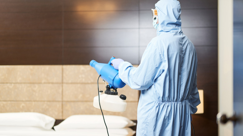 Hotel worker in hazmat gear holding an industrial sprayer of disinfectant and pointing it towards the middle of the room