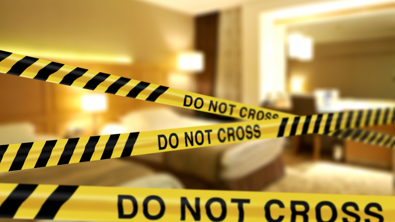 Crime scene "Do Not Cross" tape across hotel room doorway with blurred hotel room with double beds in background