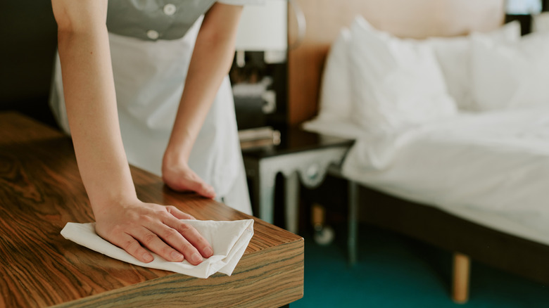 Medium close up of unrecognizable female hand using cotton napkin to dust wooden table in a hotel room with bed visible in background