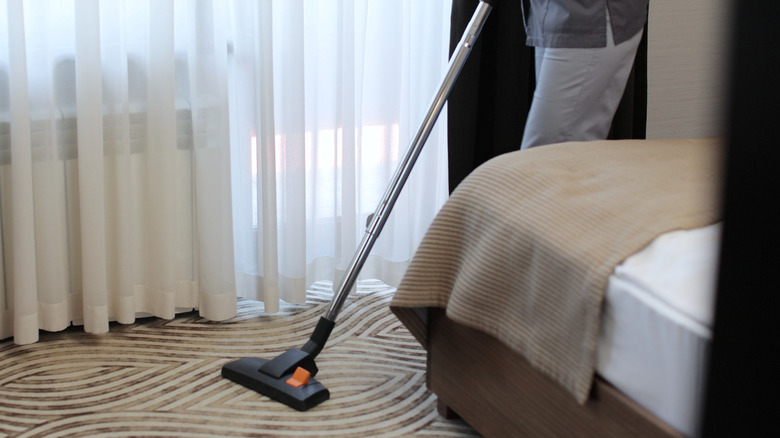 Mid-close-up of woman vacuuming a carpet in a hotel room with pants and bottom of maid uniform shirt visible