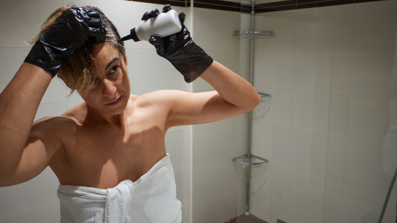 A woman with short hair dying her hair dark in a bathroom while wearing a towel.