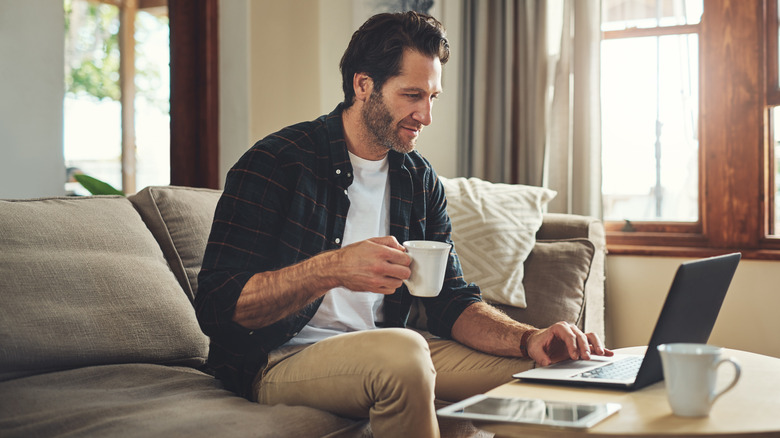 Man holding a cup of coffee while typing on laptop