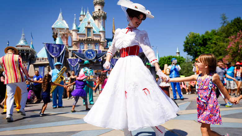 A child holding the hand of a theme park character, with a castle and park-goers in the backdrop