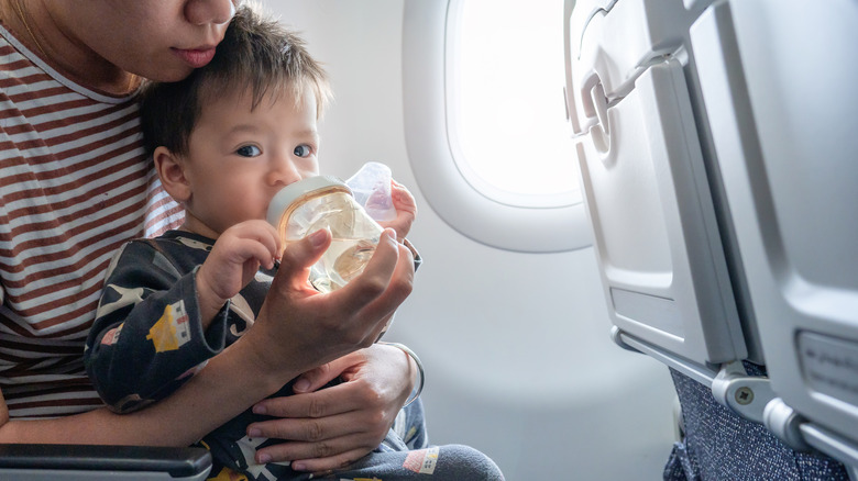A mom holds a baby with a bottle in the seat of an airplane