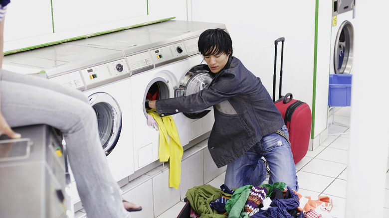 A young man with suitcase loading laundry into a laundromat machine.