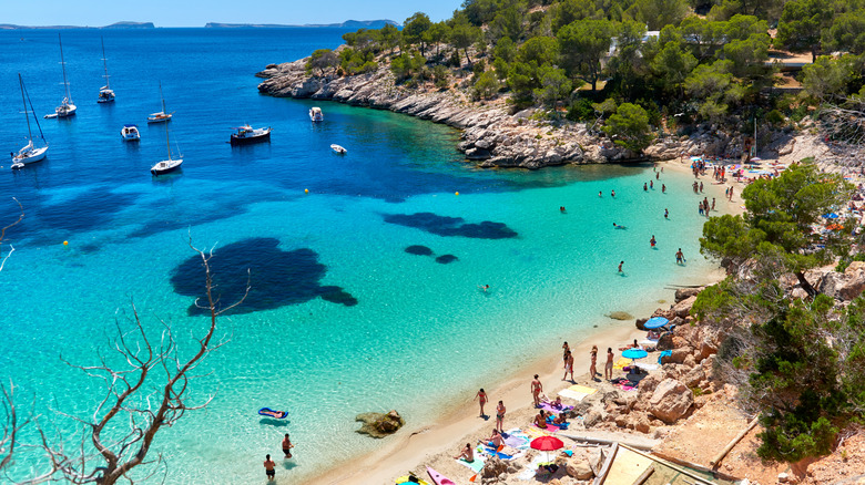 Blue water in the Cala Saldeta cove in Ibiza with yachts in the water and visitors in swimsuits on the beach