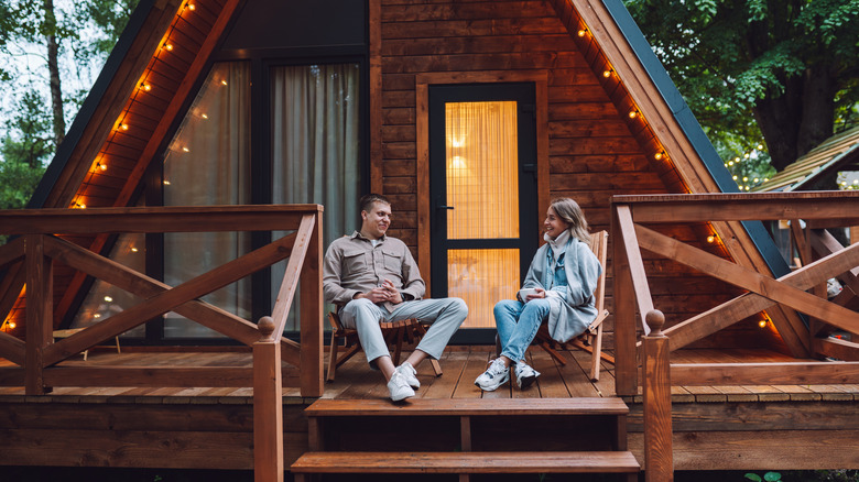 A couple relaxing on the balcony of an A-frame vacation rental in the woods.