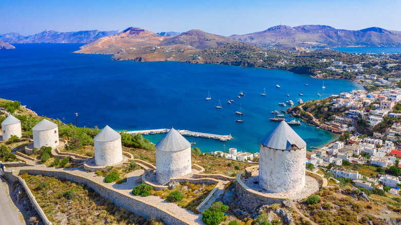 Aerial view of a row of windmills on a hill with a seafront town, sea, and mountains in the backdrop